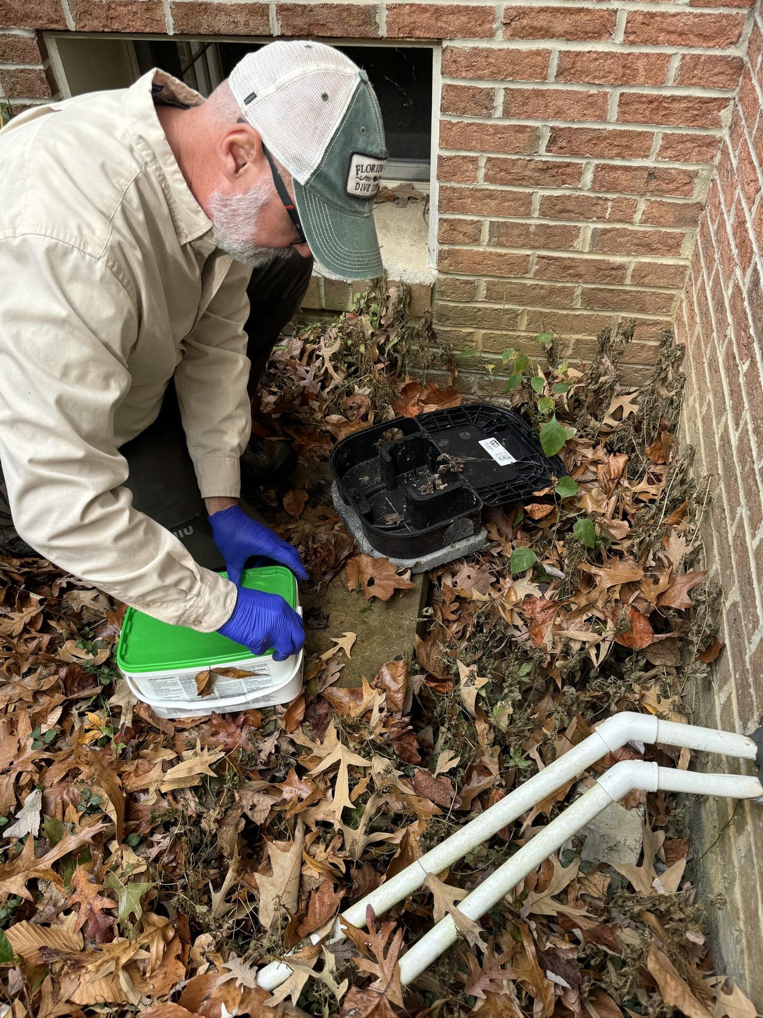 A technician inspecting a rodent bait station along a brick foundation