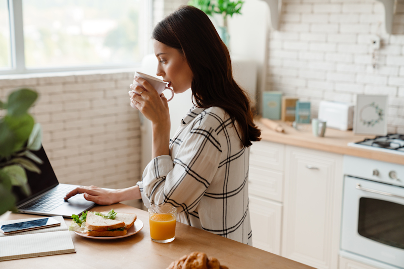 Beautiful focused woman working with laptop while having breakfast in cozy kitchen