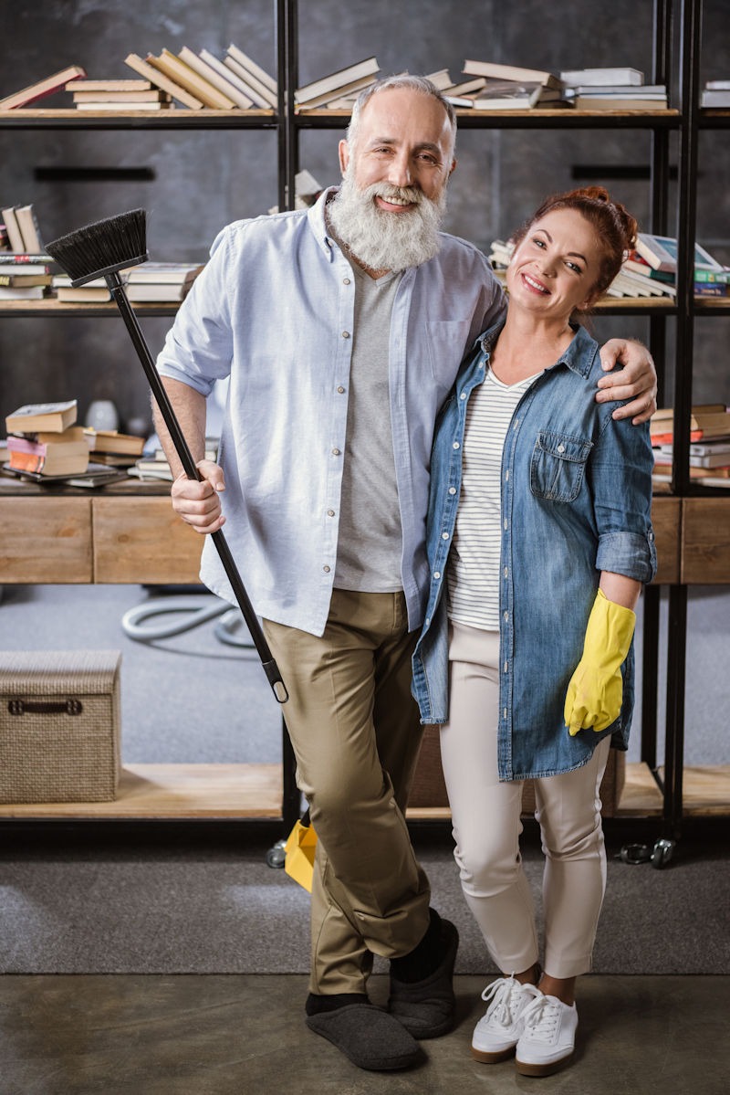 Happy mature couple standing embracing and holding broom while cleaning room together